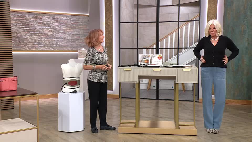 A woman in a leopard-print top gestures towards a mannequin wearing a back brace. Another woman, in a black cardigan and blue trousers, stands with her hands on her hips.