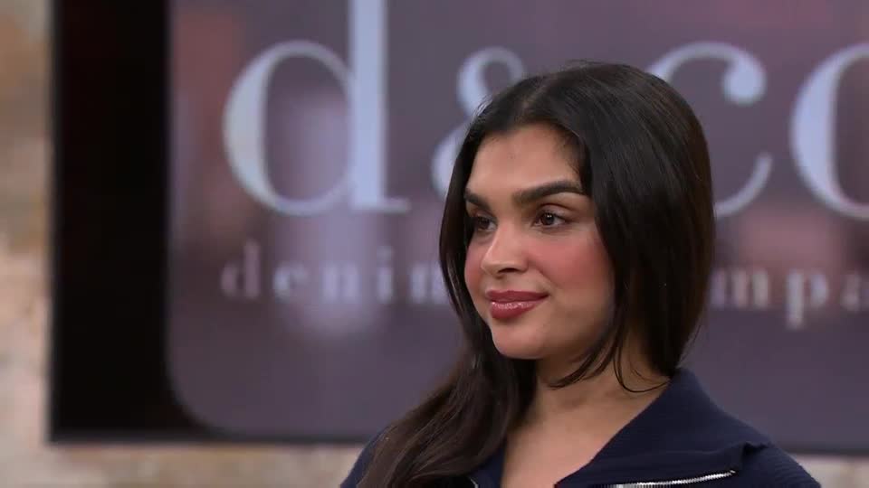 A woman with dark hair and a navy jacket smiles gently, her gaze directed slightly off-camera. Behind her, a large, stylized logo for "Denim & Co." is visible against a muted background.