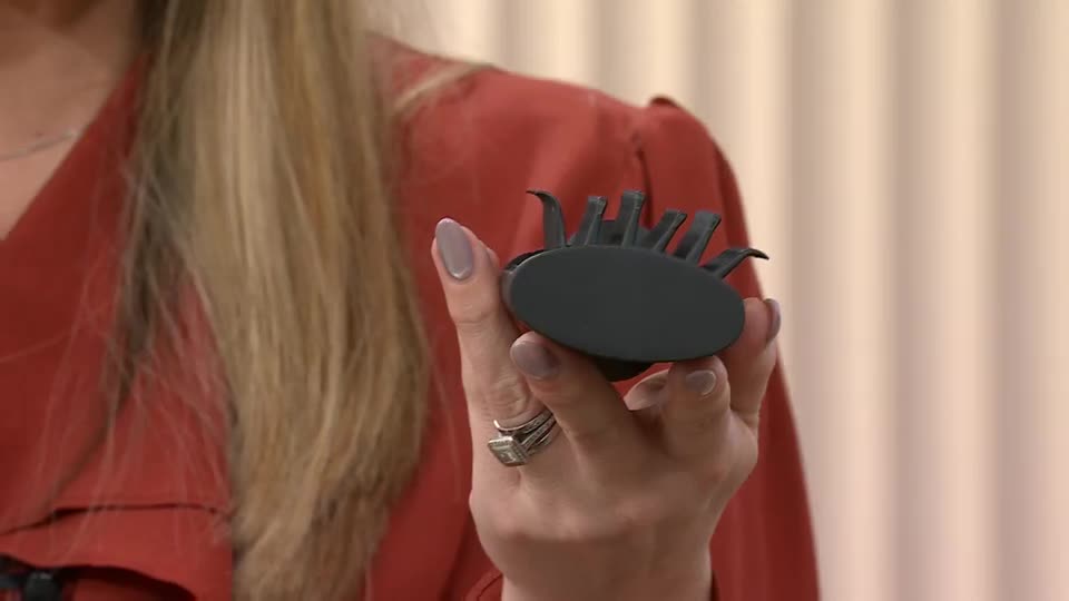 A woman on QVC holds up a black hair clip. Its oval base has several pointed prongs curving upwards.