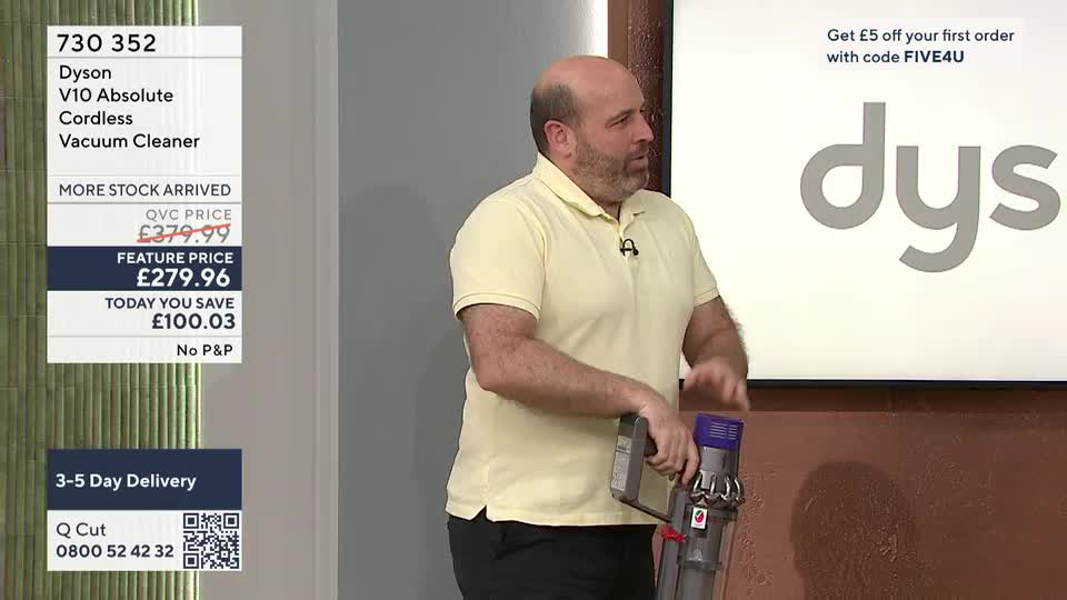 A man in a pale yellow polo shirt demonstrates a Dyson cordless vacuum cleaner. He's holding the device, its purple and grey components clearly visible.