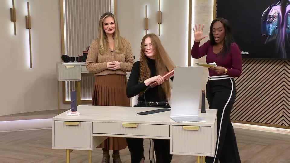 A woman with long blonde hair stands to the left, while another woman with long auburn hair straightens her own hair with a rose gold tool. A third woman, wearing a maroon top, gestures with her hand as she holds a paper.