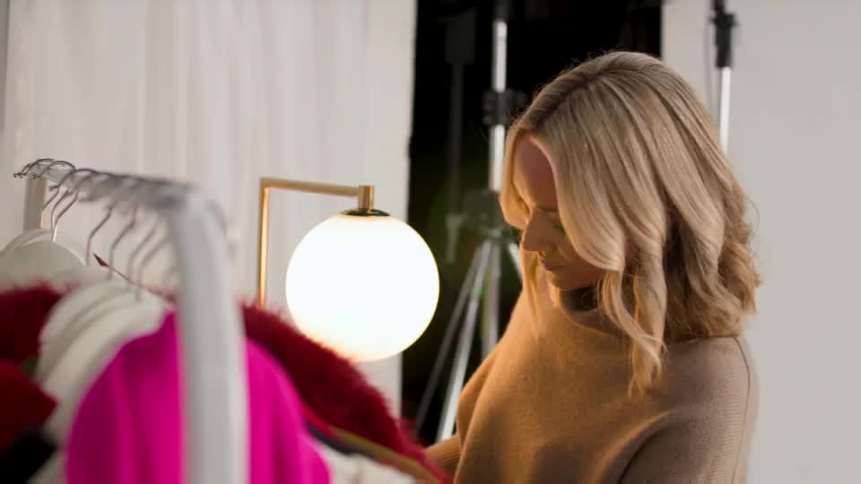 A woman with blonde hair is looking down at a rack of clothing. A bright lamp illuminates the scene, casting a warm glow on the garments.