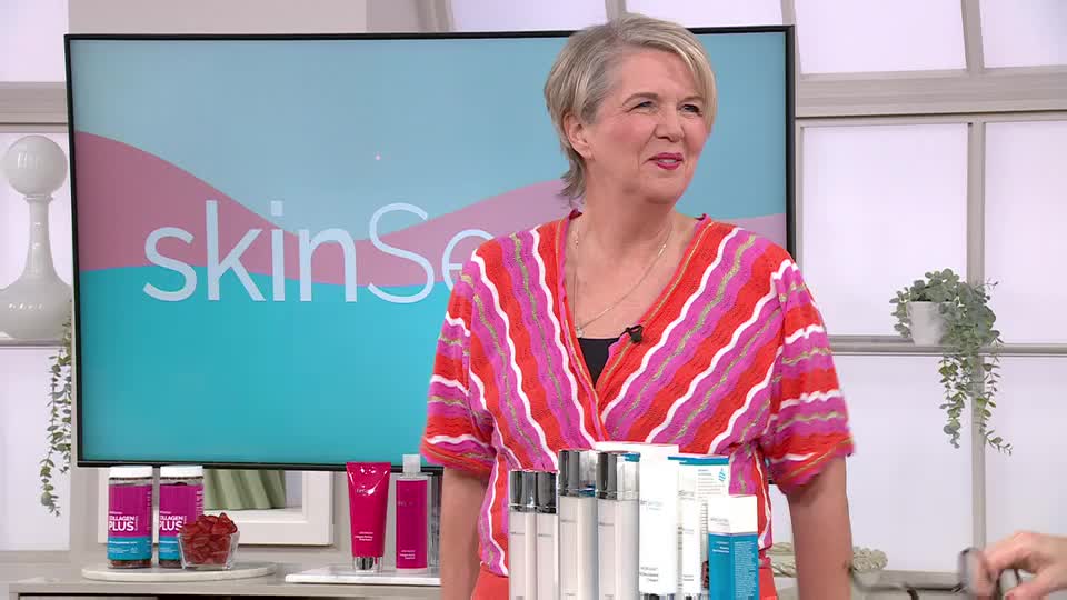 A woman in a bright pink and orange striped top smiles as she stands behind a display of skincare products. The QVC logo is visible on a screen behind her.