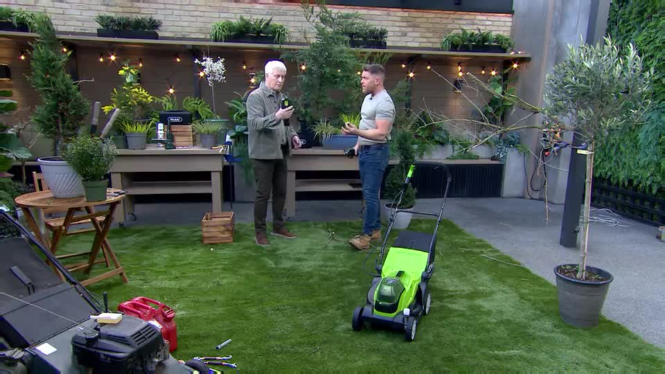 Two men stand on a patch of artificial grass, discussing gardening tools. A bright green lawnmower sits between them, ready for use. Two men stand on a patch of artificial grass, discussing gardening tools. A bright green lawnmower sits between them, ready for use.