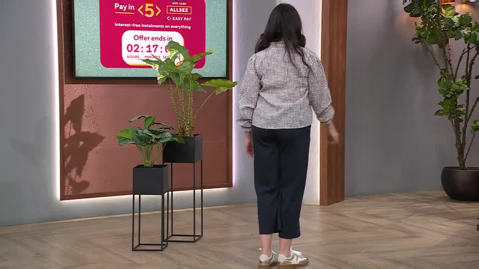 A woman in a checkered shirt and dark trousers walks away from a screen displaying a QVC offer ending soon. Two modern planters with lush green plants stand beside her. A woman in a checkered shirt and dark trousers walks away from a screen displaying a QVC offer ending soon. Two modern planters with lush green plants stand beside her.