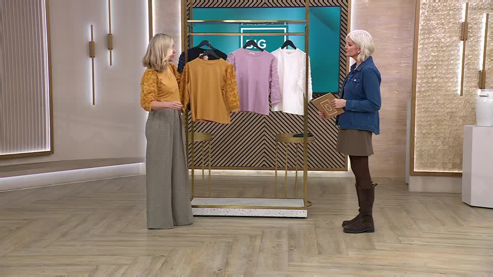 A woman in a mustard top and wide-leg trousers gestures towards a rack of jumpers. Beside her, another woman in a denim jacket holds a tablet, ready to present. A woman in a mustard top and wide-leg trousers gestures towards a rack of jumpers. Beside her, another woman in a denim jacket holds a tablet, ready to present.