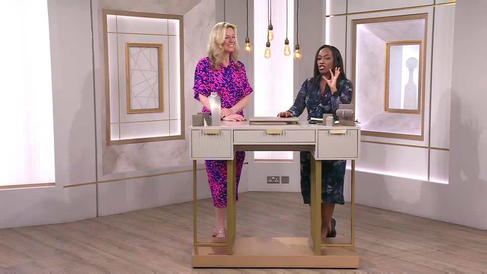 Two women stand behind a desk on a bright set, one in a vibrant pink and blue dress, the other in a darker patterned dress. The woman in the patterned dress gestures animatedly as she speaks. Two women stand behind a desk on a bright set, one in a vibrant pink and blue dress, the other in a darker patterned dress. The woman in the patterned dress gestures animatedly as she speaks.