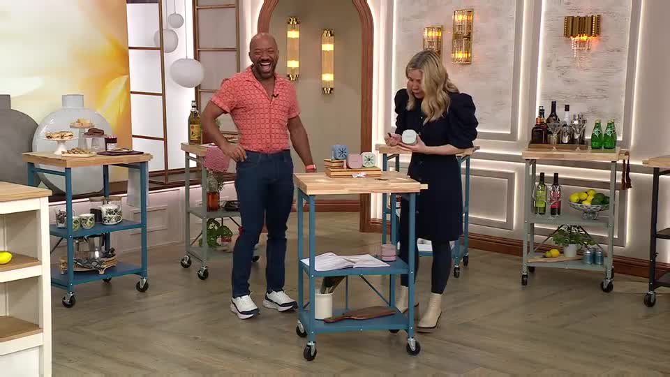 A man in a coral shirt laughs heartily as a woman in a navy dress examines a small, round object. They stand on a studio floor, surrounded by rolling carts laden with various items, suggesting a QVC presentation. A man in a coral shirt laughs heartily as a woman in a navy dress examines a small, round object. They stand on a studio floor, surrounded by rolling carts laden with various items, suggesting a QVC presentation.
