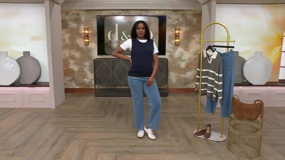 A woman models a navy sleeveless top over a white t-shirt and wide-leg jeans. On a nearby rack, a striped sweater and more jeans are displayed. A woman models a navy sleeveless top over a white t-shirt and wide-leg jeans. On a nearby rack, a striped sweater and more jeans are displayed.
