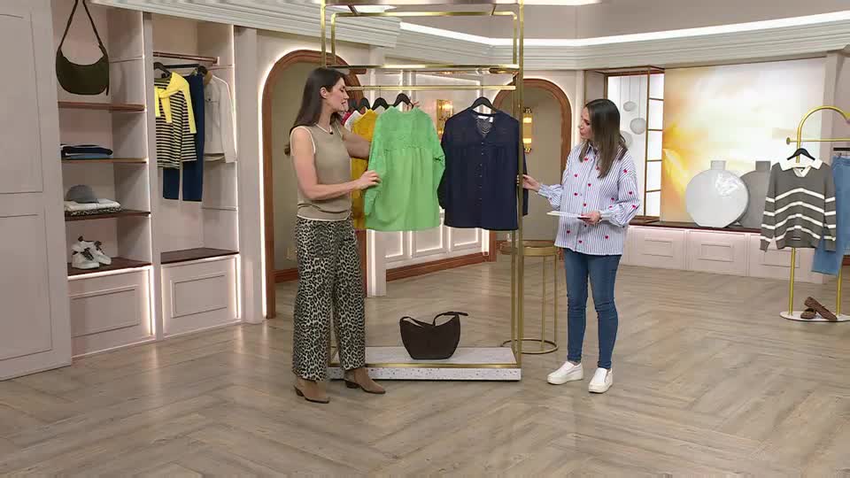 A woman in leopard-print trousers holds up a bright green top. Beside her, another woman in a striped shirt gestures towards a dark blue blouse. A woman in leopard-print trousers holds up a bright green top. Beside her, another woman in a striped shirt gestures towards a dark blue blouse.