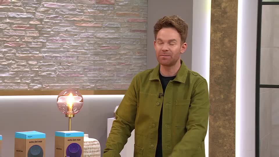 A man in a green jacket stands on a QVC set, gesturing as he speaks. Behind him, a decorative lamp glows warmly next to boxes for an Echo Dot. A man in a green jacket stands on a QVC set, gesturing as he speaks. Behind him, a decorative lamp glows warmly next to boxes for an Echo Dot.