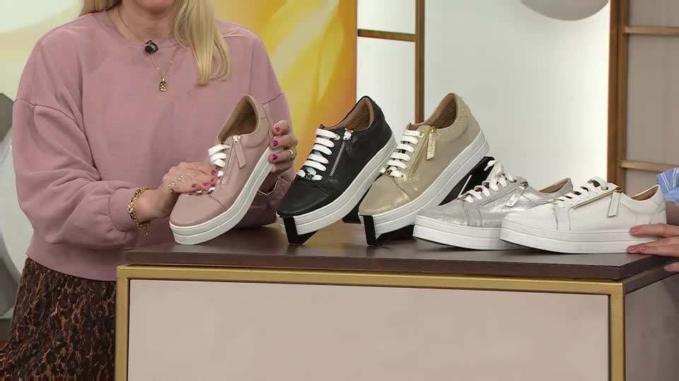 A woman in a pink sweatshirt holds up a blush pink sneaker, showcasing its white laces and side zipper. Next to it, a row of similar sneakers in black, gold, silver, and white are displayed on a tiered stand. A woman in a pink sweatshirt holds up a blush pink sneaker, showcasing its white laces and side zipper. Next to it, a row of similar sneakers in black, gold, silver, and white are displayed on a tiered stand.
