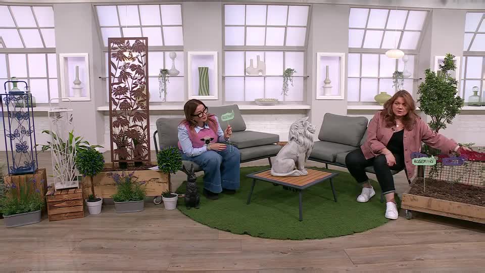 A woman in a pink jacket gestures towards a raised garden bed, while another woman in a blue top and pink vest holds up a green sign. A woman in a pink jacket gestures towards a raised garden bed, while another woman in a blue top and pink vest holds up a green sign.