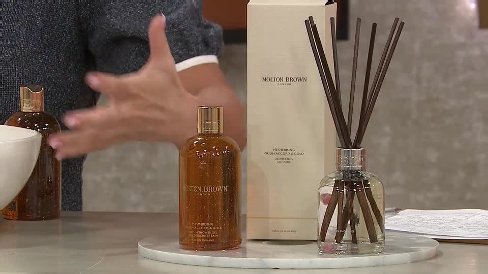 A woman's hands gesture enthusiastically as she presents Molton Brown products, a diffuser with dark reeds standing tall beside a bottle of amber liquid. The scene unfolds on a QVC broadcast, showcasing luxury bath and body items from the UK brand. A woman's hands gesture enthusiastically as she presents Molton Brown products, a diffuser with dark reeds standing tall beside a bottle of amber liquid. The scene unfolds on a QVC broadcast, showcasing luxury bath and body items from the UK brand.