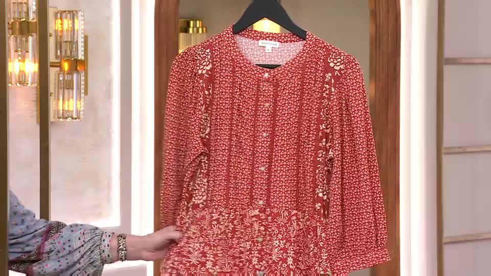 A hand reaches out to adjust the hem of a red, patterned dress hanging on a rack. The dress is displayed against a backdrop of warm lighting and decorative wall sconces, typical of a QVC presentation. A hand reaches out to adjust the hem of a red, patterned dress hanging on a rack. The dress is displayed against a backdrop of warm lighting and decorative wall sconces, typical of a QVC presentation.