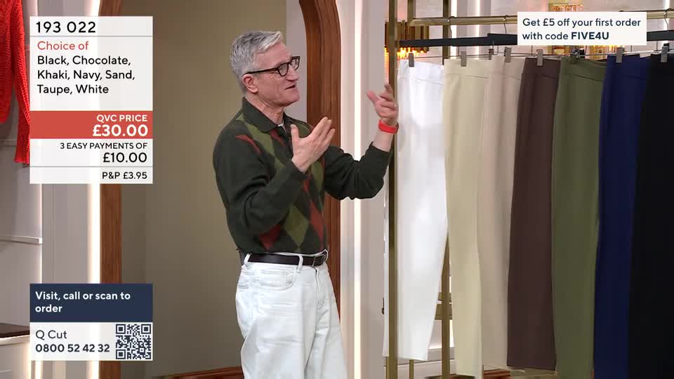 A man in a patterned jumper gestures towards a rack of neatly hung trousers in various colours. The QVC price of £30.00 is displayed prominently on a screen beside him. A man in a patterned jumper gestures towards a rack of neatly hung trousers in various colours. The QVC price of £30.00 is displayed prominently on a screen beside him.