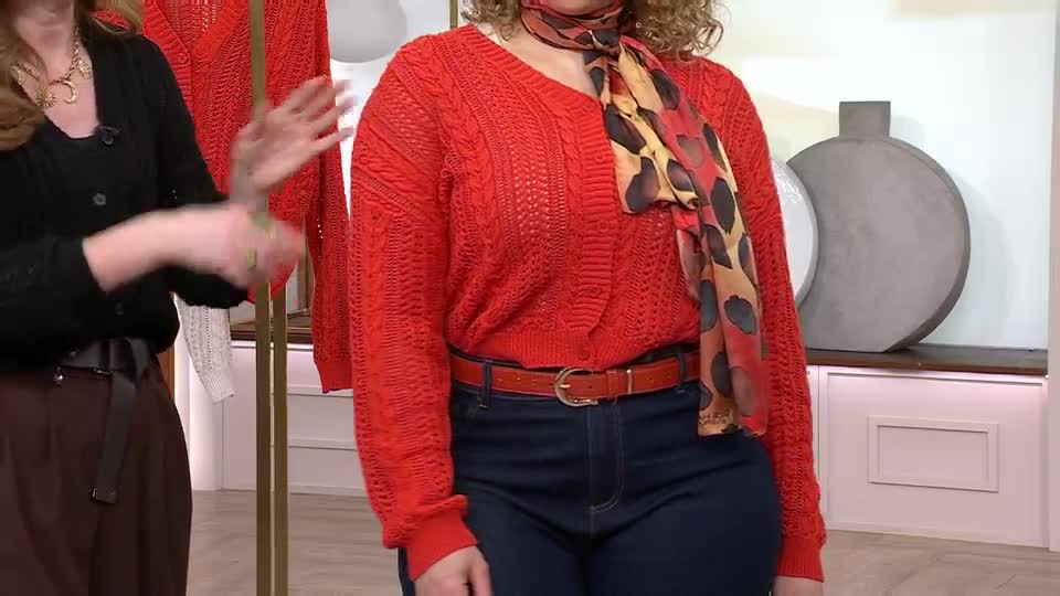 A woman in a black cardigan gestures with her hands as a model in a vibrant red cable-knit sweater stands beside her. The model wears a patterned scarf and a brown belt over dark jeans, presenting a QVC fashion segment. A woman in a black cardigan gestures with her hands as a model in a vibrant red cable-knit sweater stands beside her. The model wears a patterned scarf and a brown belt over dark jeans, presenting a QVC fashion segment.