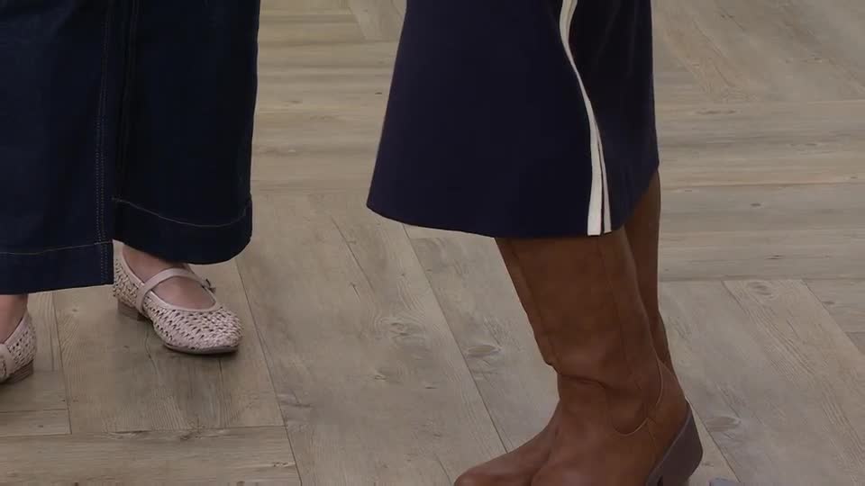 A woman in a pale pink, woven flat shoe stands on a wooden floor. Beside her, another woman's legs are clad in dark blue trousers and a brown boot. A woman in a pale pink, woven flat shoe stands on a wooden floor. Beside her, another woman's legs are clad in dark blue trousers and a brown boot.