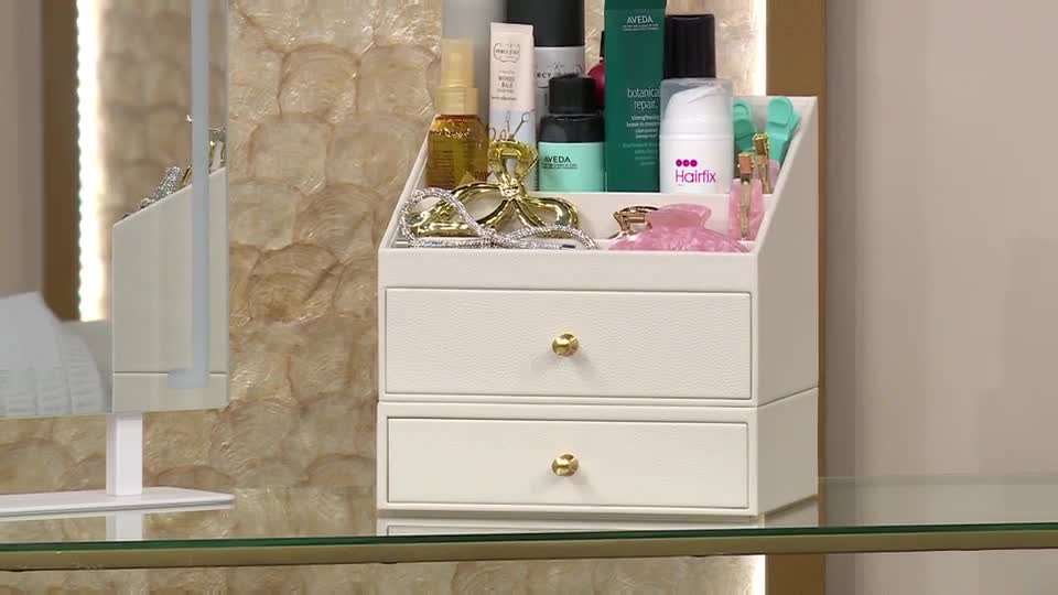 A white organiser, filled with various hair products and accessories, sits on a glass shelf. The QVC presenter is about to showcase these items. A white organiser, filled with various hair products and accessories, sits on a glass shelf. The QVC presenter is about to showcase these items.