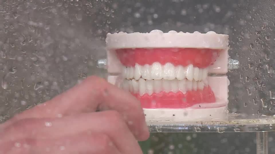 A hand reaches towards a set of dentures, which are being sprayed with water. The water droplets cling to the clear plastic casing, obscuring the background.
