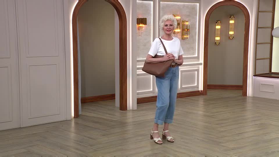 A woman stands on a polished wooden floor, holding a brown handbag. She's wearing a white t-shirt and light blue jeans, with her hair styled in short, curly blonde.