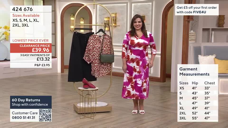 A woman in a vibrant pink and white floral dress stands on a QVC set. She gestures to a rack of clothing, showcasing a patterned blouse and black trousers.