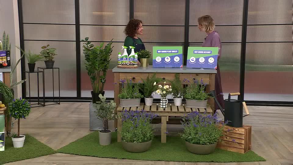 Two women stand behind a wooden display table laden with plants and gardening products. A black watering can rests on a wooden crate to the right.