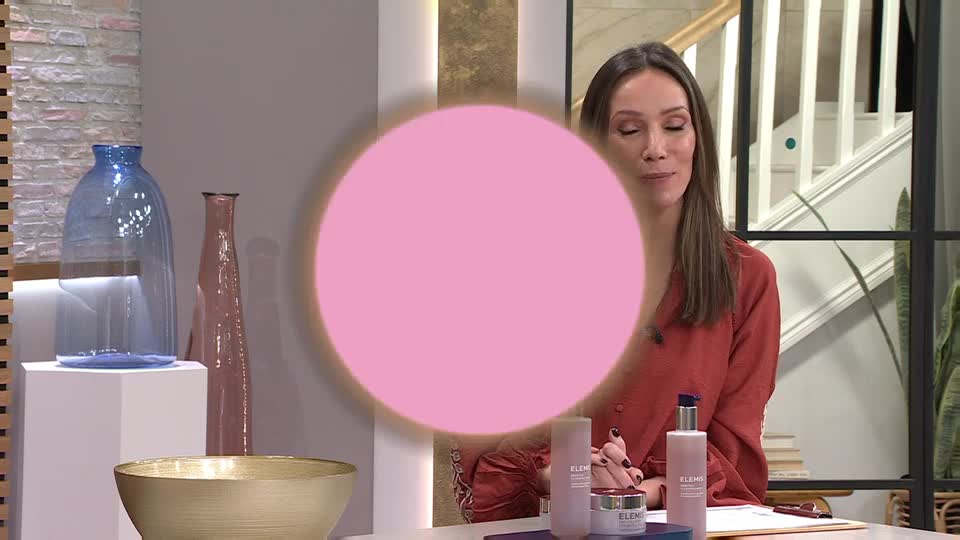 A woman in a rust-coloured top is speaking, gesturing with her hands near a display of skincare products. Behind her, a large pink circle hovers, obscuring part of the set.