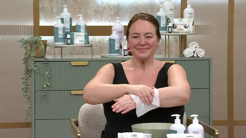 A woman on QVC is wiping her hands with a white cloth, smiling. Behind her, rows of skincare bottles are neatly arranged on shelves.