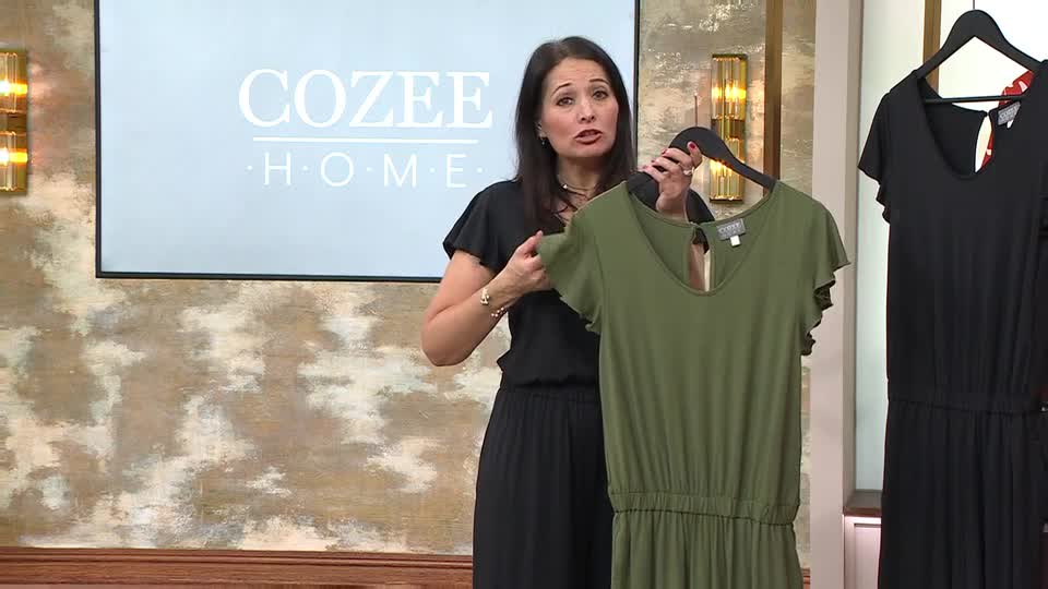 A woman on QVC is presenting a green jumpsuit, holding it up by the hanger. Another black jumpsuit hangs beside it. A woman on QVC is presenting a green jumpsuit, holding it up by the hanger. Another black jumpsuit hangs beside it.