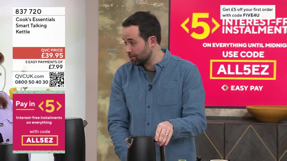 A man in a blue shirt gestures towards a black kettle. Behind him, a bright pink screen advertises interest-free instalments on QVC. A man in a blue shirt gestures towards a black kettle. Behind him, a bright pink screen advertises interest-free instalments on QVC.