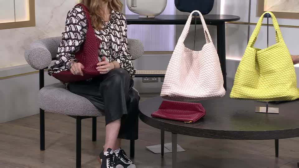 A woman in a patterned blouse and black trousers holds a deep red woven tote bag. On the table before her sit a pale pink tote and a bright yellow tote, both also woven, along with a matching red clutch. A woman in a patterned blouse and black trousers holds a deep red woven tote bag. On the table before her sit a pale pink tote and a bright yellow tote, both also woven, along with a matching red clutch.