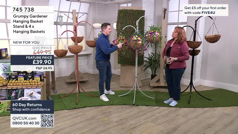 A man in a blue jumper gestures towards a display of hanging baskets on a stand. A woman in a pink jacket stands nearby, holding a small silver cup. A man in a blue jumper gestures towards a display of hanging baskets on a stand. A woman in a pink jacket stands nearby, holding a small silver cup.