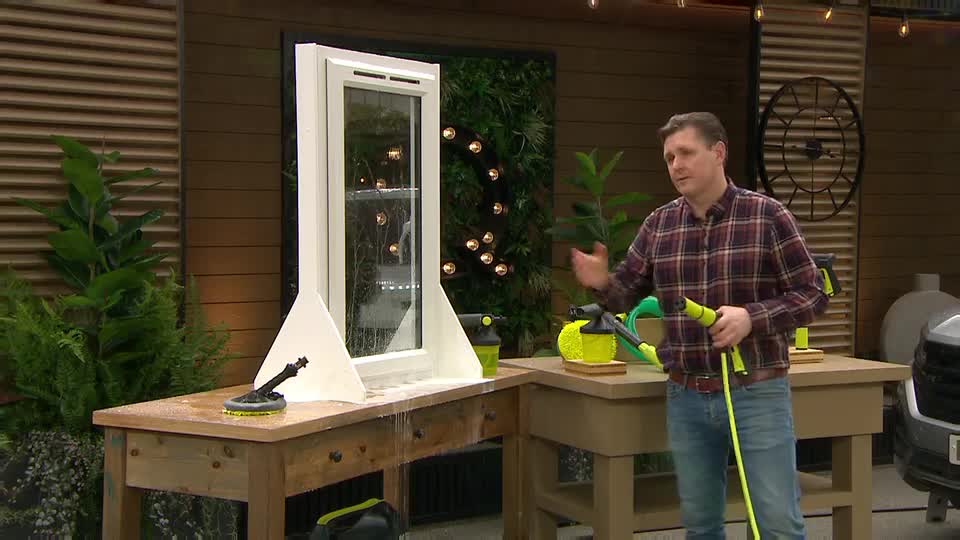 A man in a plaid shirt holds a yellow and green hose, demonstrating a cleaning product. Water sprays from the hose onto a white frame and a wooden table. A man in a plaid shirt holds a yellow and green hose, demonstrating a cleaning product. Water sprays from the hose onto a white frame and a wooden table.