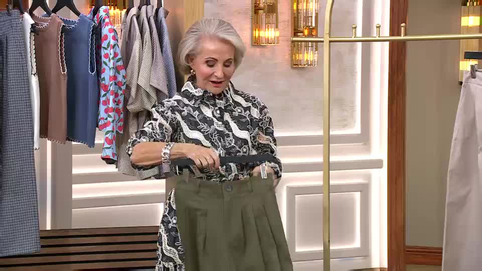 A woman on QVC holds up a pair of olive green trousers. Behind her, a rack of clothes hangs, including a dress with a cherry print. A woman on QVC holds up a pair of olive green trousers. Behind her, a rack of clothes hangs, including a dress with a cherry print.