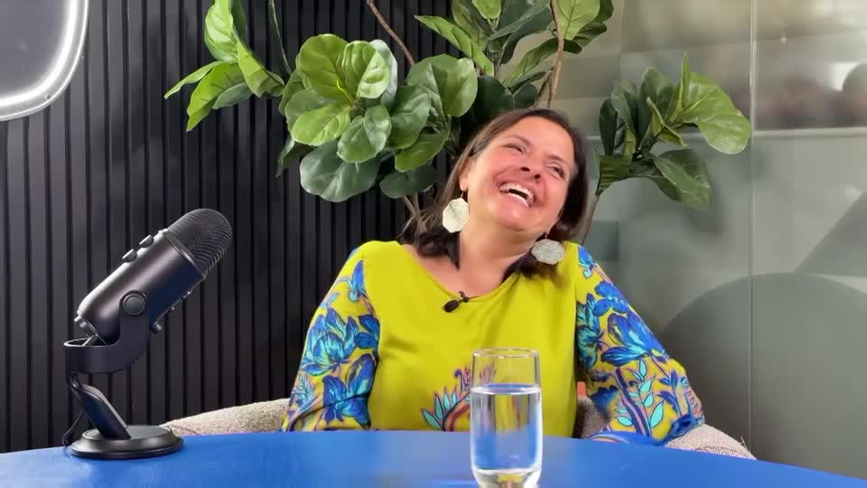 A woman in a bright yellow top with blue floral sleeves throws her head back in laughter. A microphone sits to her left, and a glass of water is in front of her.