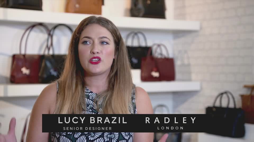 A woman with bright red lipstick speaks animatedly in front of shelves displaying Radley handbags. She's a designer for the London-based company, likely discussing their latest collection as seen on QVC.