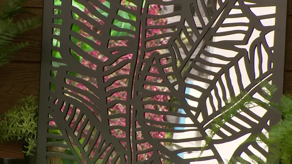 A dark metal screen, cut with a fern leaf pattern, stands in front of a display of vibrant pink flowers. Beside it, a lush green plant adds a touch of natural texture.