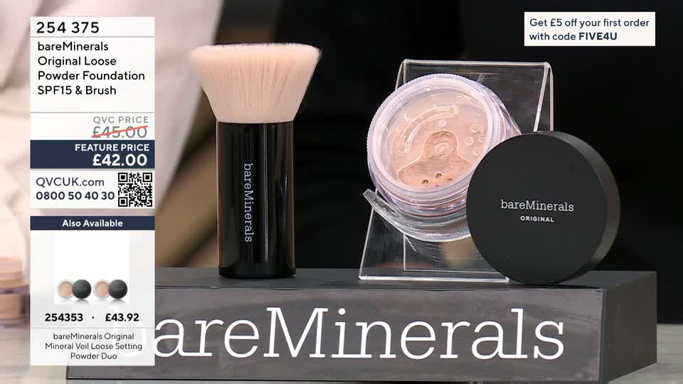 A large, fluffy brush with a black handle stands next to an open jar of bareMinerals powder. The jar is propped on a clear stand, and a closed black container of the same product sits beside it.