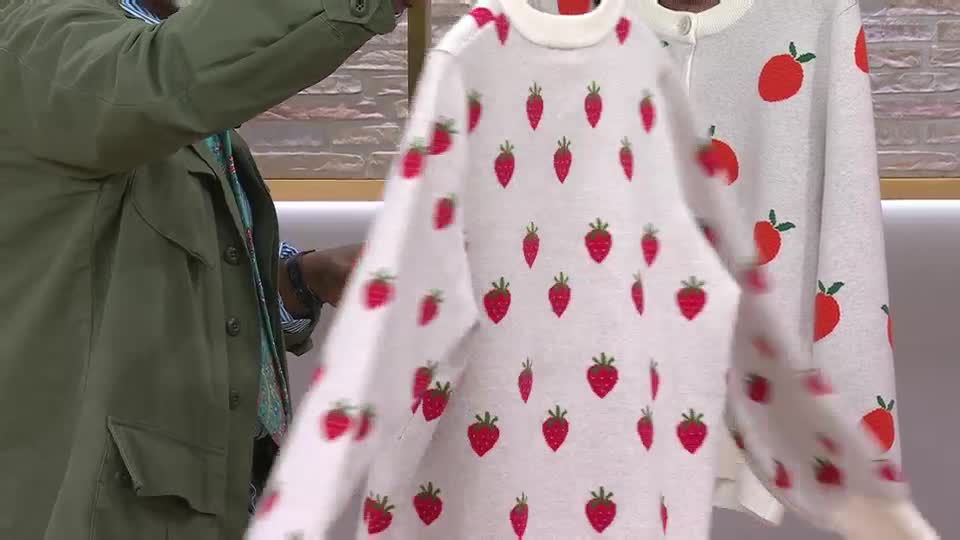 Someone's holding up a white jumper covered in little red strawberries. Another jumper with larger strawberries hangs next to it. Someone's holding up a white jumper covered in little red strawberries. Another jumper with larger strawberries hangs next to it.