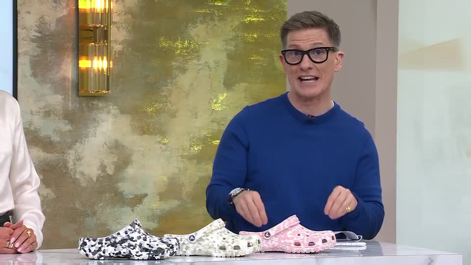 A man in a blue sweater gestures enthusiastically towards three pairs of patterned Crocs. The shoes, in black and white, beige, and pink, sit on a counter as he speaks. A man in a blue sweater gestures enthusiastically towards three pairs of patterned Crocs. The shoes, in black and white, beige, and pink, sit on a counter as he speaks.