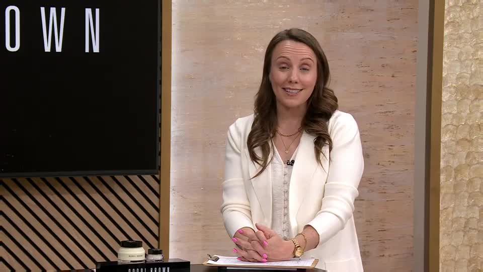 A woman in a cream blazer smiles, her hands clasped in front of her. Beside her, two jars of Bobbi Brown skincare sit on a dark tray, ready for their QVC presentation. A woman in a cream blazer smiles, her hands clasped in front of her. Beside her, two jars of Bobbi Brown skincare sit on a dark tray, ready for their QVC presentation.