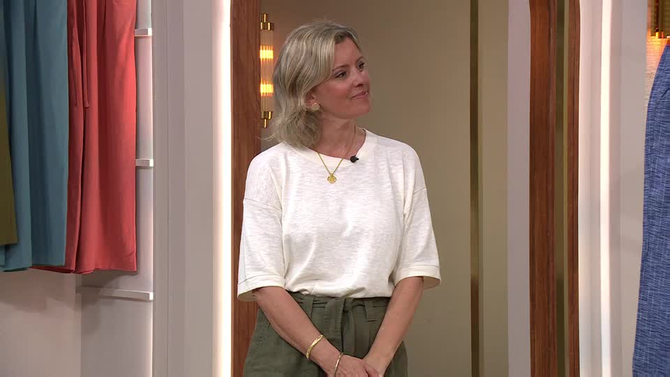 A woman stands in front of a rack of colourful clothes, likely on QVC. She smiles as she looks slightly off-camera, her hands clasped at her waist. A woman stands in front of a rack of colourful clothes, likely on QVC. She smiles as she looks slightly off-camera, her hands clasped at her waist.