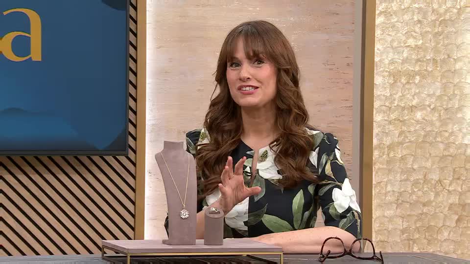 A presenter on QVC in the UK gestures with her hands as she talks about a diamond pendant and ring displayed on stands. The backdrop behind her features a textured wall and a screen showing a graphic. A presenter on QVC in the UK gestures with her hands as she talks about a diamond pendant and ring displayed on stands. The backdrop behind her features a textured wall and a screen showing a graphic.
