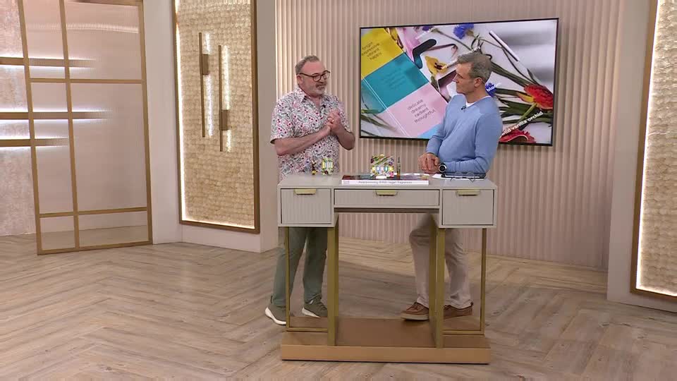 Two men stand on a studio floor, one in a patterned shirt, the other in a blue jumper. A large screen behind them displays colourful books and flowers. Two men stand on a studio floor, one in a patterned shirt, the other in a blue jumper. A large screen behind them displays colourful books and flowers.