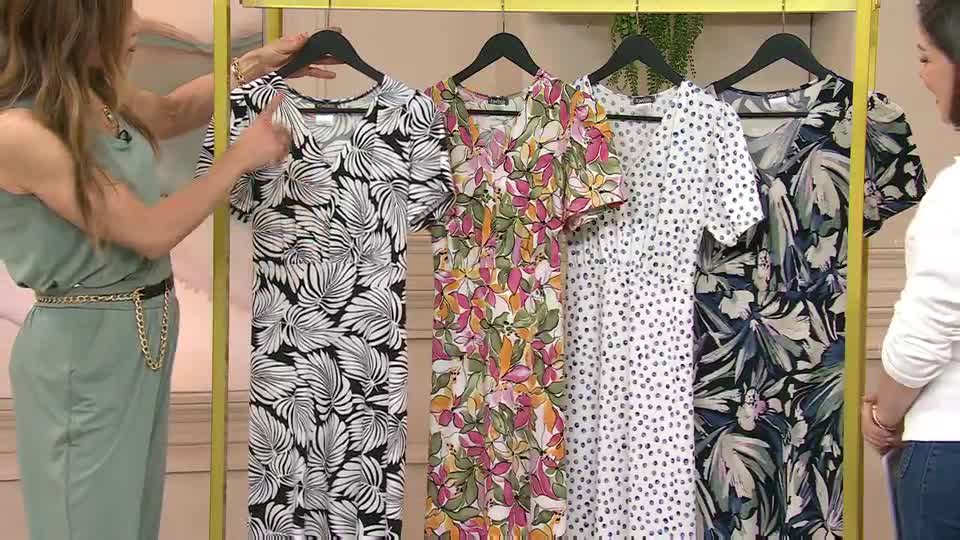 A woman in a pale green jumpsuit gestures towards a rack of dresses. The dresses, displayed on black hangers, feature bold floral and leaf prints. A woman in a pale green jumpsuit gestures towards a rack of dresses. The dresses, displayed on black hangers, feature bold floral and leaf prints.