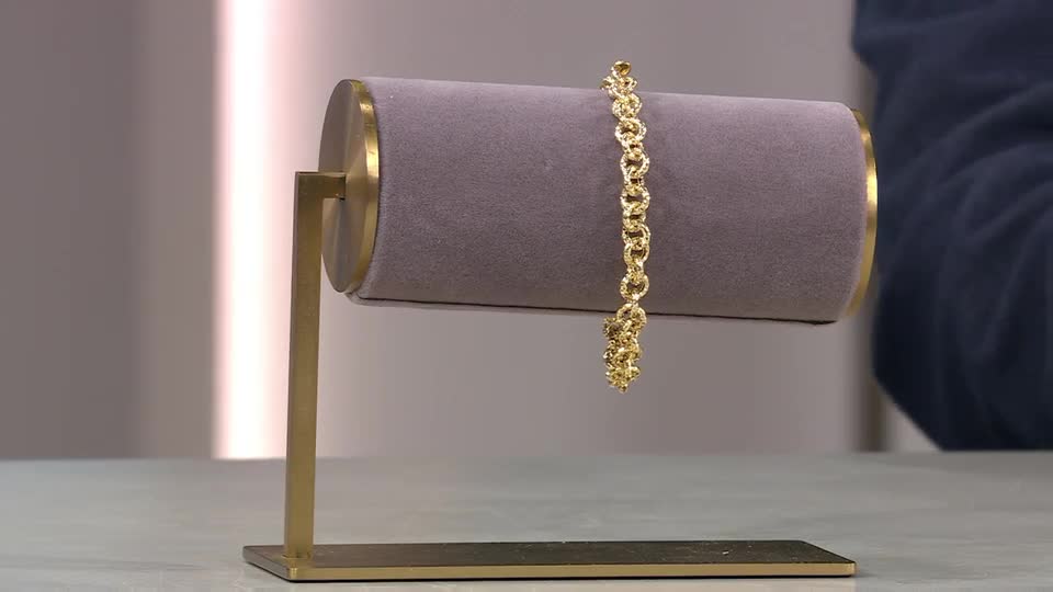 A gold bracelet hangs from a grey velvet display. A hand in a dark blue sleeve reaches towards it. A gold bracelet hangs from a grey velvet display. A hand in a dark blue sleeve reaches towards it.