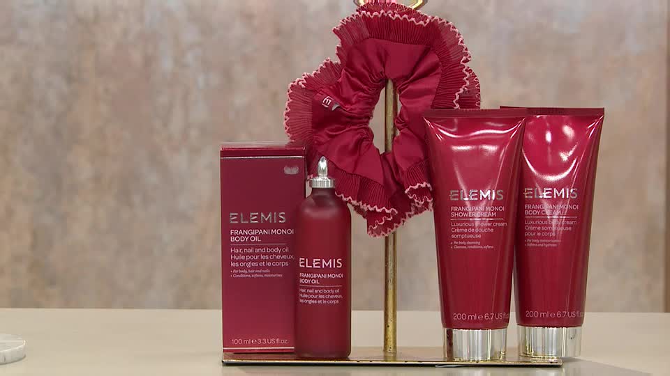 A display of Elemis products, including body oil and creams, is presented on a gold stand. A large, ruffled red scrunchie sits behind the bottles, catching the light.