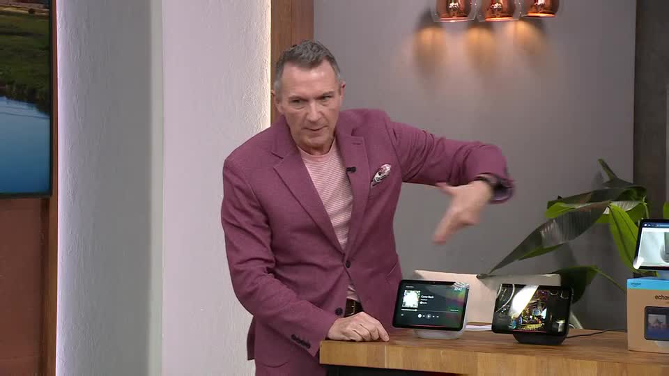 A man in a vibrant pink suit gestures animatedly towards a row of smart displays on a wooden counter. He's mid-sentence, likely explaining a product to viewers watching QVC in the UK.