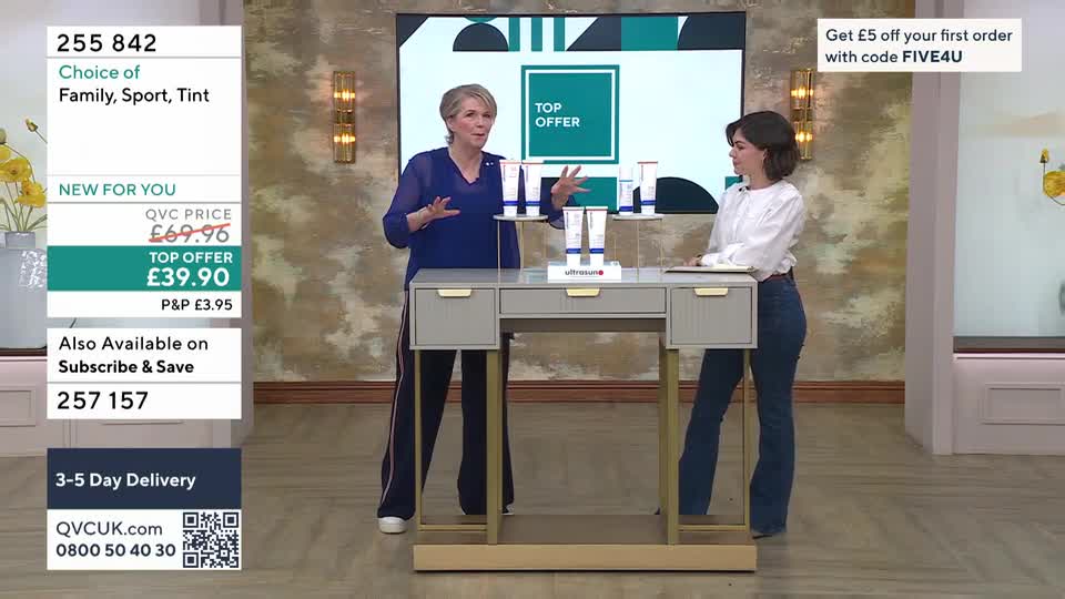 A woman in a blue top gestures enthusiastically towards a display of skincare products on a grey desk. Beside her, another woman in a white shirt and jeans listens intently.